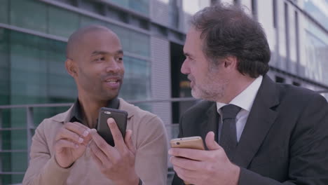 Professional-men-having-discussion-on-meeting-with-smartphones-near-office-building